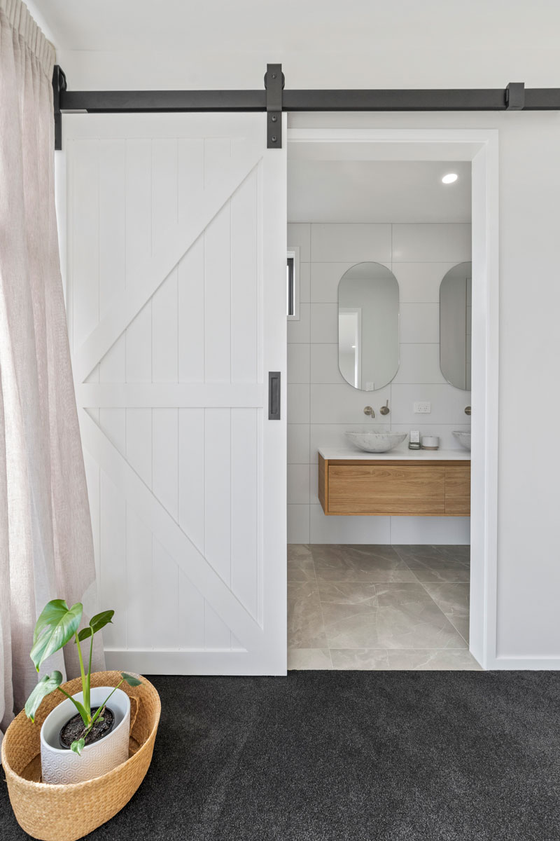 barn door to ensuite with timber New Tech vanity and oval mirrors in designer home